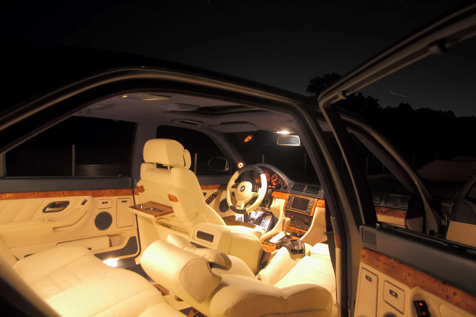 BMW E38 cabin glowing under interior lights at night, cream leather and burl wood seen through open doors beneath a starry sky